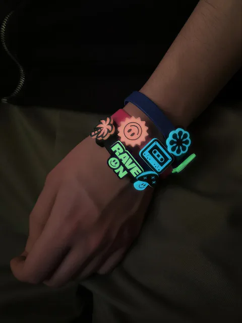 Kilka bransoletek FLUO na nadgarstku ze świecącymi przypinkami: RAVE ON, palma, buźka, kwiatek i kaseta