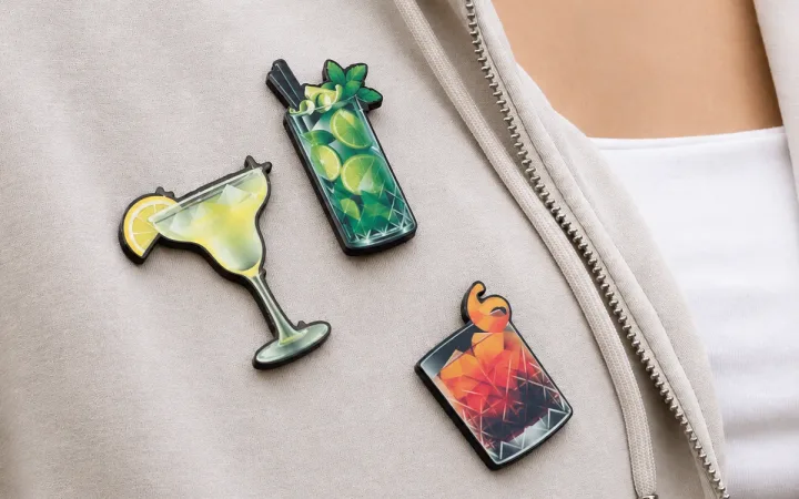 Trzy piny w kształcie drinków – margarita, mojito i old fashioned – przypięte do bluzy